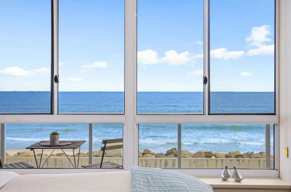Large windows invite natural light and ocean breezes into the bedroom, offering uninterrupted views of the sea. A small outdoor balcony is visible with a table and two chairs, creating a serene spot to enjoy the coastal landscape.