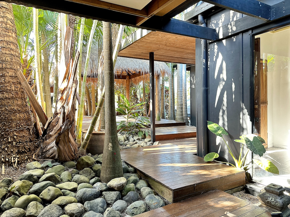 A tranquil outdoor space features wooden decking surrounded by lush tropical plants and smooth stones. The area is shaded by palm trees and includes a thatched-roof structure in the background, creating a serene atmosphere for relaxation.