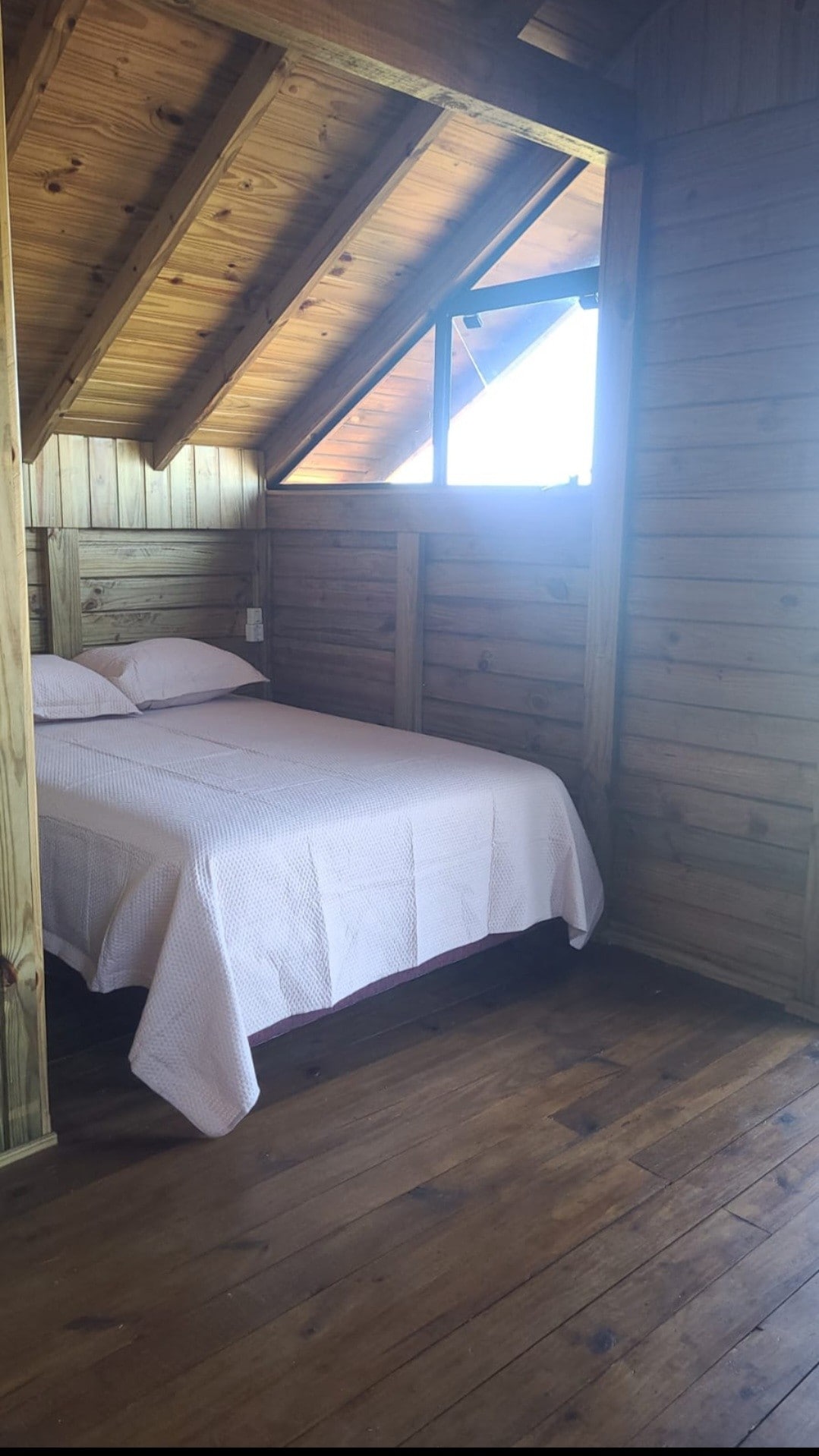 A cozy sleeping area features a spacious bed draped with a light-colored quilt. Natural wood paneling lines the walls, and a small window allows natural light to fill the room, providing a warm and inviting atmosphere.