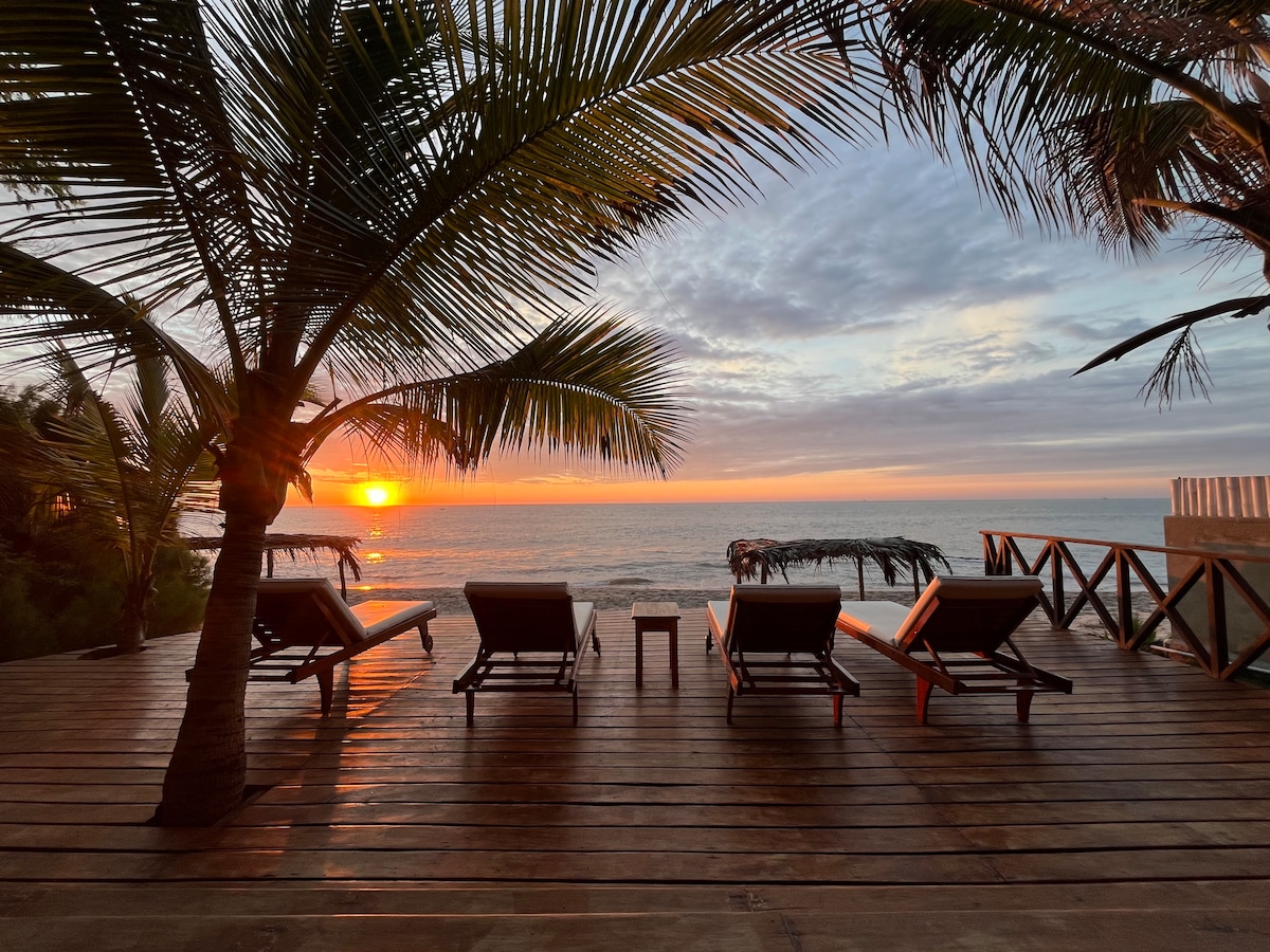 A private beach front deck features three lounge chairs positioned to face the ocean, where a sunset casts warm hues across the sky. Palm trees frame the scene, providing a tropical atmosphere as the gentle waves lap at the shore.