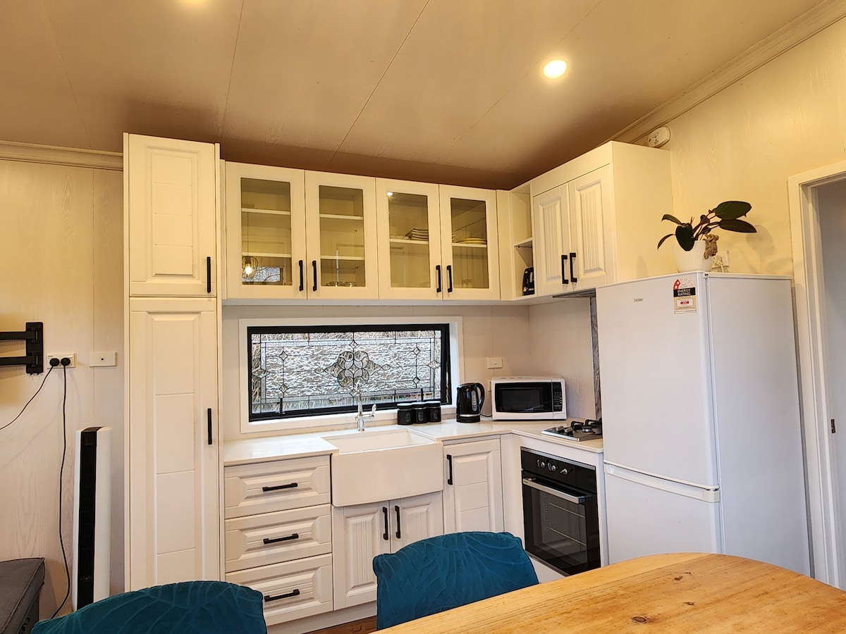 A compact kitchen area features white cabinetry and a large window above a farmhouse sink. Modern appliances include a microwave, refrigerator, and oven. A coffee maker is present on the countertop, with two teal chairs visible at the dining table nearby.