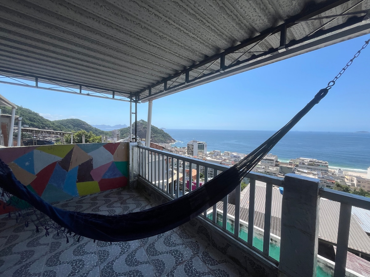 A spacious rooftop terrace features a hammock, providing sweeping views of the ocean and Copacabana beach. Colorful wall murals add vibrancy to the space, while the clear blue sky enhances the tranquil atmosphere. The layout invites relaxation amidst the scenic surroundings.