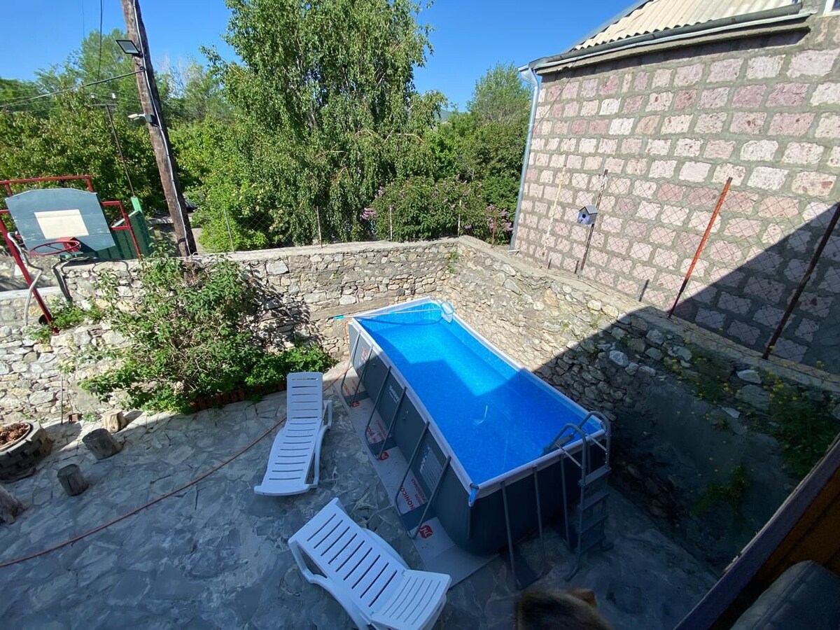 An outdoor area features a rectangular above-ground pool with clear blue water, surrounded by a stone wall. Four white lounge chairs are positioned nearby on a stone patio, with greenery visible in the background, including trees and a basketball hoop.
