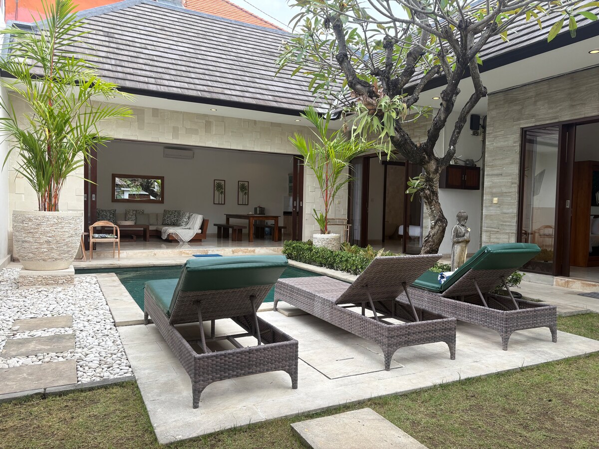 Two outdoor sun loungers are positioned beside a private pool surrounded by landscaped gardens and tropical plants. The villa's living area is visible in the background, featuring large glass doors that invite natural light. A decorative statue is placed among the greenery.