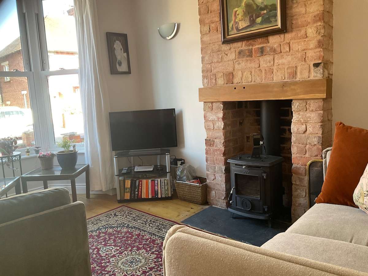 The lounge area features a brick fireplace with a woodburner, complemented by a flat-screen television. A soft rug lies on the wooden floor, and comfortable seating options are present, along with a decorative wall art piece. Natural light is allowed through the windows covered with light curtains.