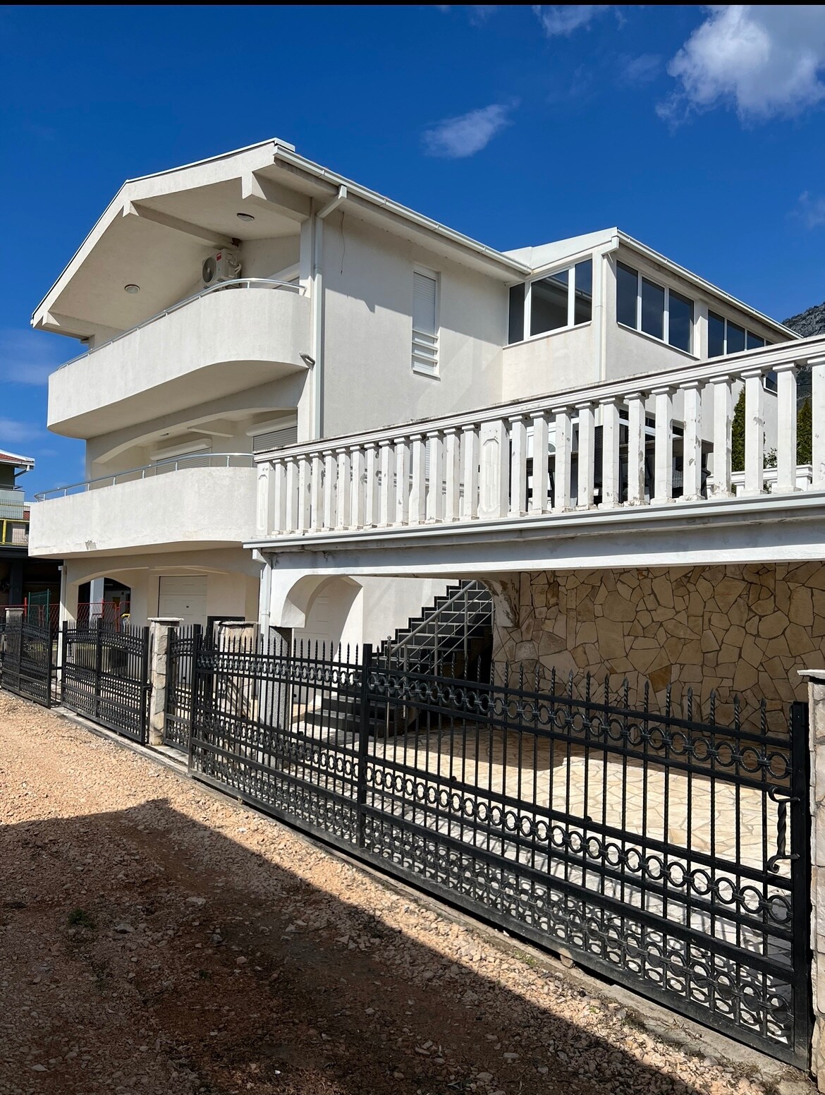 A modern two-story building is situated under a clear blue sky. The exterior features a white facade, multiple balconies with railings, and stone accents. A wrought iron fence surrounds the property, providing a sense of privacy. Stairs lead to the entrance, framed by landscaped surroundings.