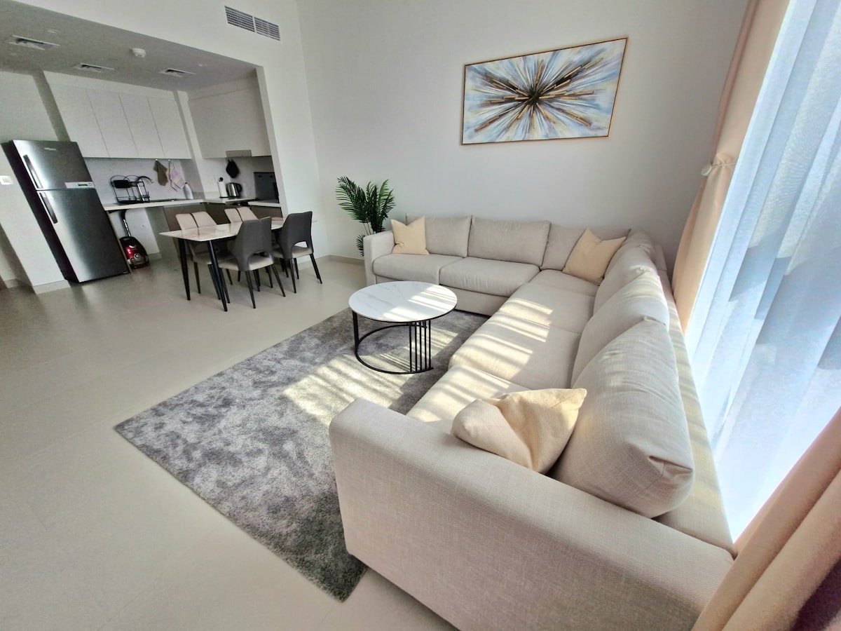 A spacious living area is presented with a light-colored sectional sofa and a round coffee table set on a soft area rug. Nearby, a dining table is visible, accompanied by dark chairs. Natural light filters through large windows, creating a bright and inviting ambiance.