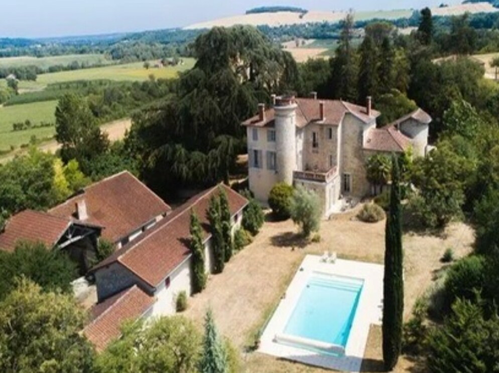An aerial view captures a charming house resembling a small château, surrounded by lush greenery and gardens. A clear swimming pool is visible in the foreground, with sun loungers arranged nearby. The expansive outdoor space features mature trees and an inviting landscape.