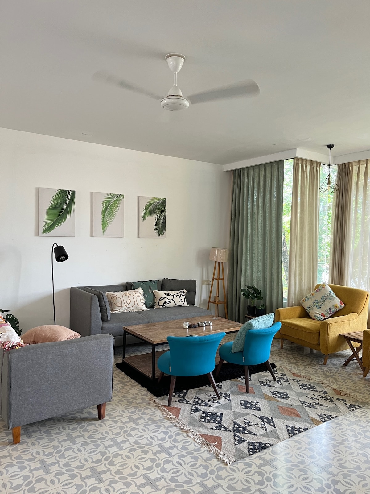 The living area features a mix of modern and cozy furnishings, including plush sofas and colorful accent chairs. Natural light enters through large windows, illuminating the space. Decorative wall art adds a touch of greenery, while a patterned rug enhances the ambiance.