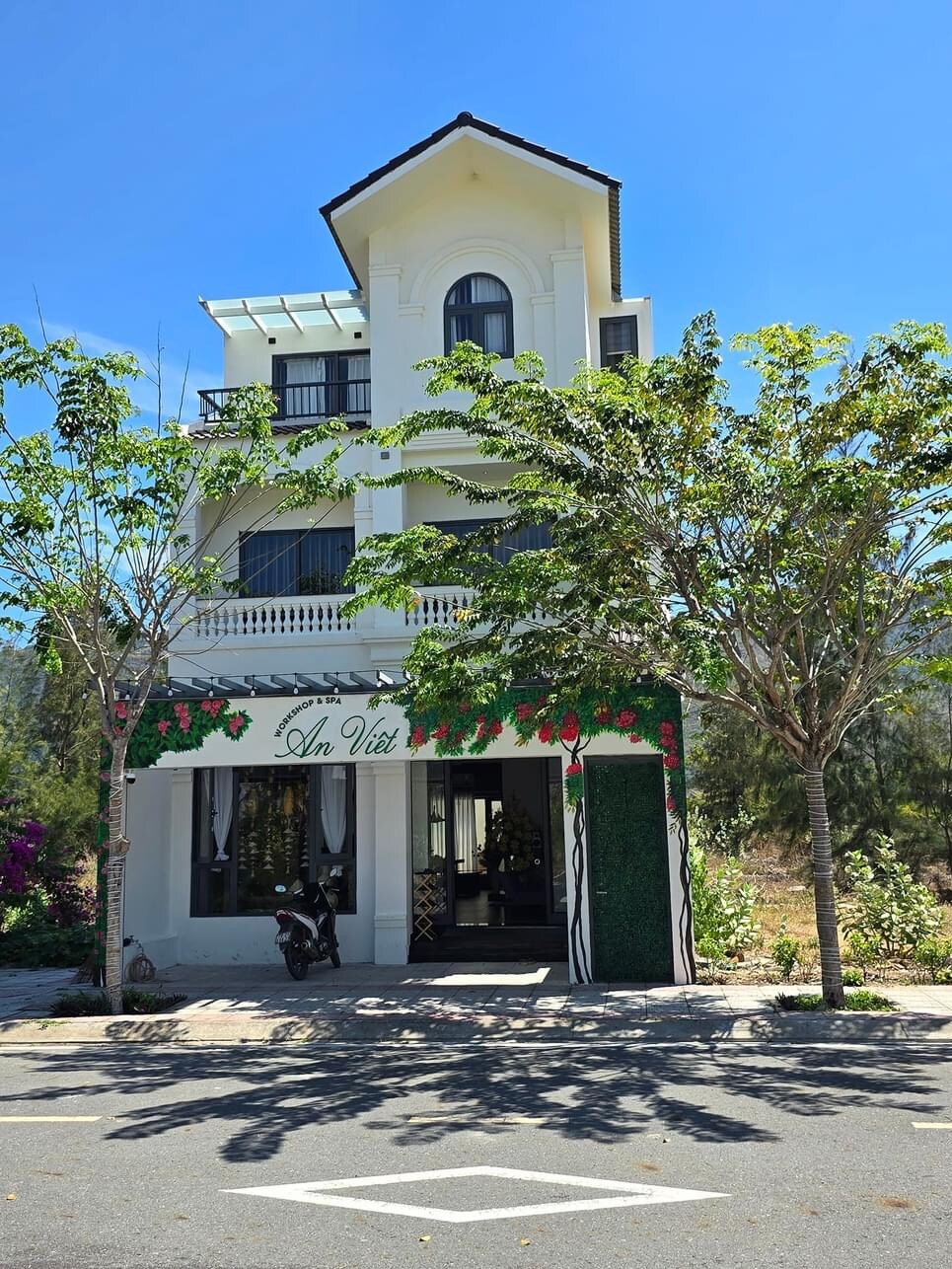 A three-story building is featured, with a white façade and decorative floral accents framing the entrance. Tall trees provide shade in front, while a clear blue sky enhances the overall appearance of the location. A subtly welcoming atmosphere is presented by the greenery.