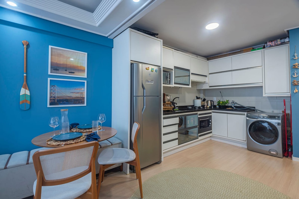 A kitchen featuring modern appliances, including a refrigerator and a washing machine, is complemented by a dining area. A round table with two chairs is positioned against a blue accent wall adorned with framed photographs, adding a touch of color to the space.
