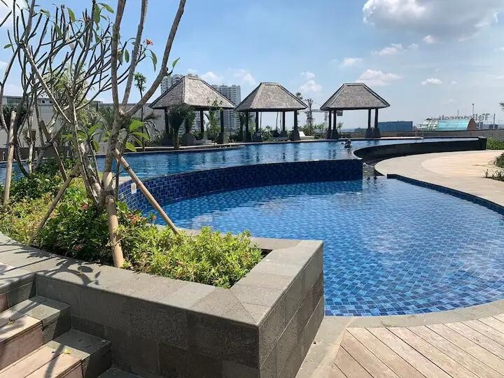 An outdoor swimming pool features vibrant blue tiles and is bordered by lush greenery. Thatch-roofed pavilions provide shaded seating around the pool area, enhancing the tranquil environment. The sun shines brightly overhead, creating a welcoming atmosphere for relaxation.