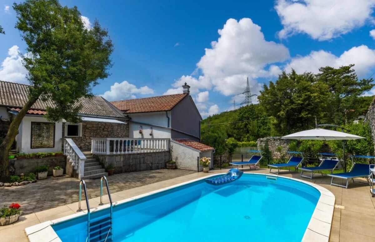 An outdoor area features a private swimming pool surrounded by a spacious terrace. Sun loungers are arranged nearby under large umbrellas, offering shaded seating. Lush greenery and a stone building add to the tranquil setting, while a clear blue sky enhances the inviting atmosphere.