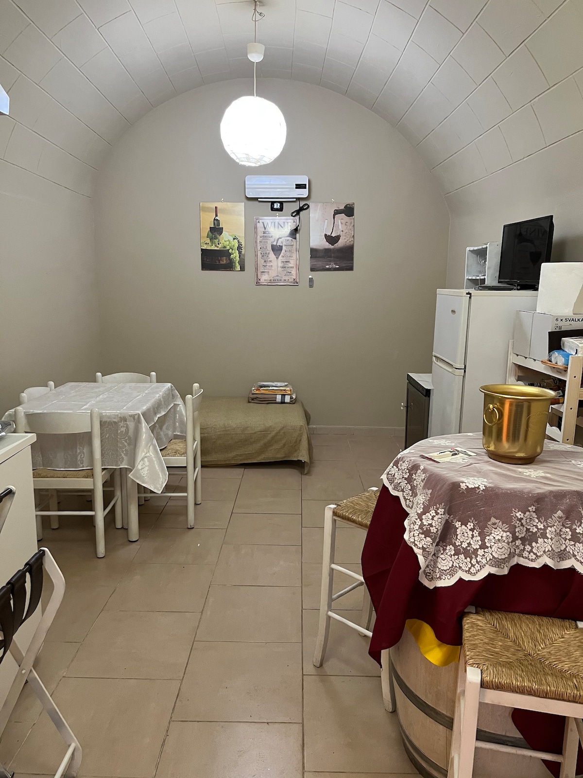 An arched room features whitewashed walls and tiled floors. A dining area includes two small tables with light-colored tablecloths. A vintage barrel serves as a table base. Kitchen appliances, including a refrigerator and a microwave, are visible against the wall. Local artwork adorns the back wall.