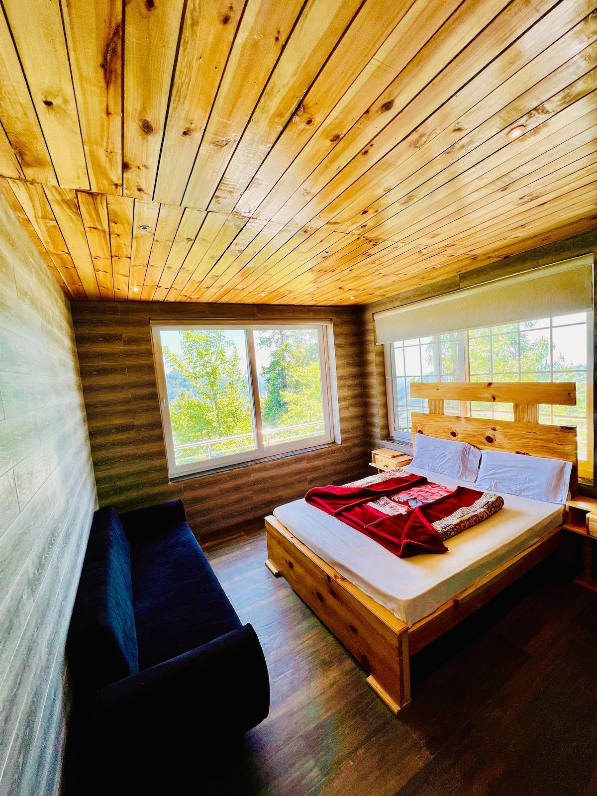 A cozy bedroom features a wooden ceiling and walls, allowing natural light to fill the space through large windows. A spacious bed with a colorful throw blanket is complemented by a small sofa, creating a welcoming and relaxed atmosphere.