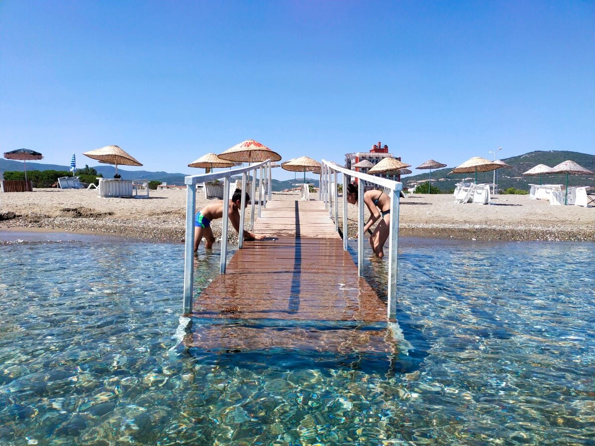 A wooden pier extends over clear water, providing access to a sandy beach. Sun umbrellas are positioned along the shoreline, offering shade. The serene landscape includes distant hills and crystal clear water, inviting relaxation and leisure activities.