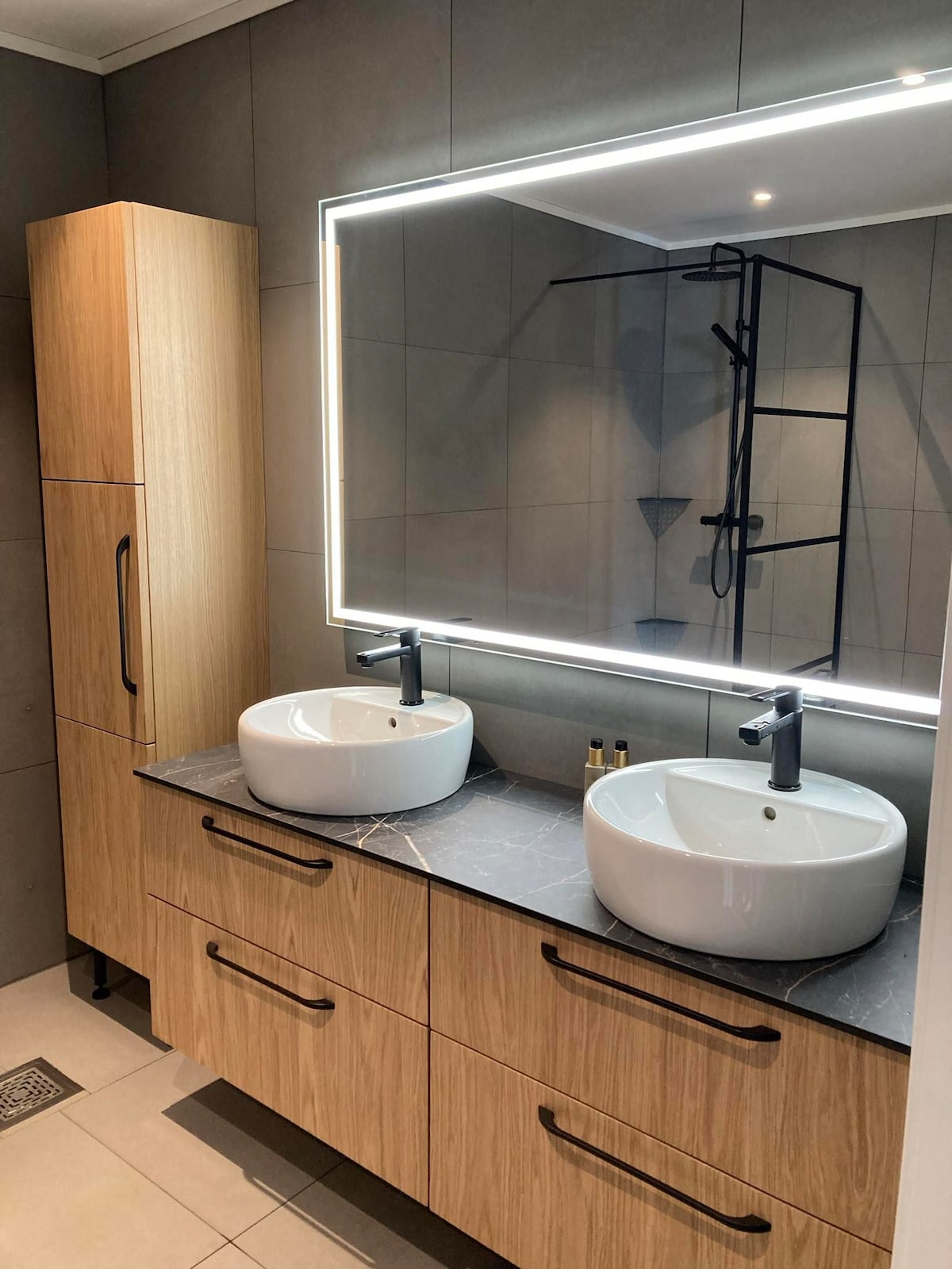 A modern bathroom is featured, showcasing a wooden vanity with two round sinks placed on a dark countertop. A large mirror with integrated lighting is mounted above the sinks. A sleek shower area is visible behind the vanity, enhancing the room's contemporary design.