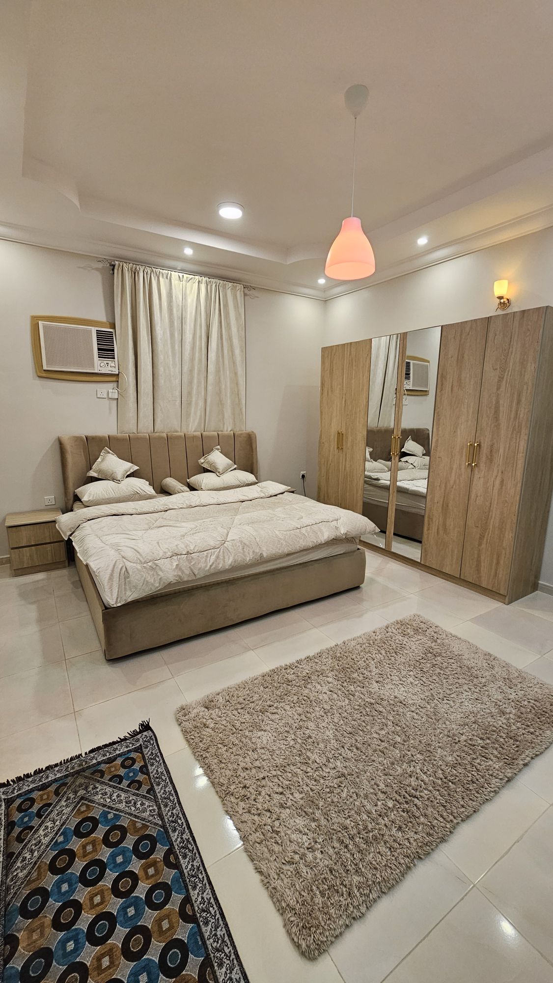 A well-appointed bedroom featuring a spacious bed with light bedding. The room includes a stylish wardrobe with sliding doors and a fluffy area rug on the floor. Natural light enters through a window adorned with soft curtains, while a wall-mounted air conditioner is visible.