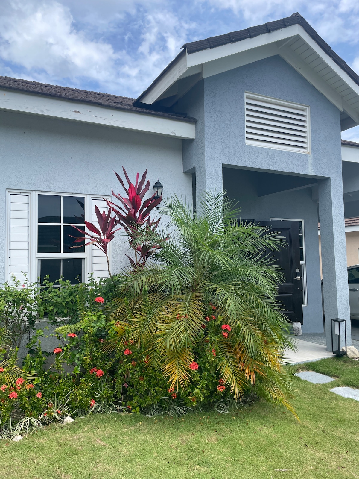 The entrance of the home is framed by vibrant tropical plants, including red and green foliage, and colorful flowers. A well-manicured lawn leads to the black front door, complemented by soft lighting fixtures on either side. The house features a modern design with white shutters.