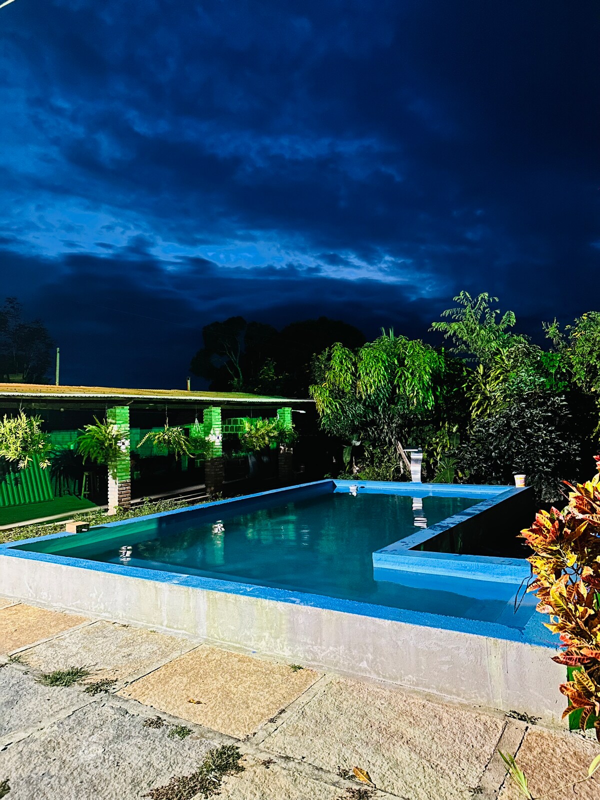 A tranquil pool area is surrounded by lush greenery, with lights reflecting off the water's surface. The sky is filled with deep blue clouds, creating a serene ambiance in the evening light. The pool features a simple rectangular design and is framed by stone pathways.