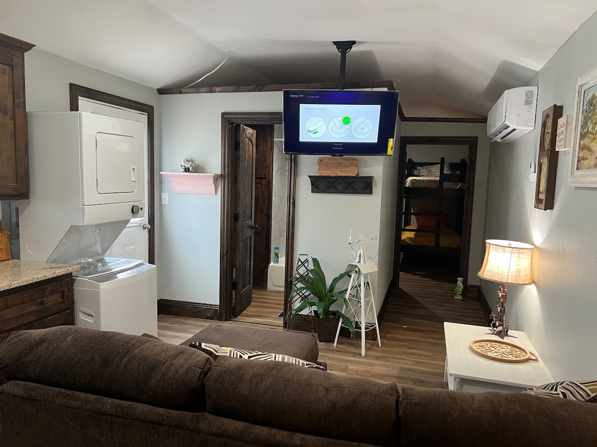 A spacious living area includes a comfortable sofa, with a mounted television above a laundry unit. A small table with decorative elements is positioned beside a lamp. A bunk bed is visible in the adjacent area, along with a small indoor plant placed on the floor.