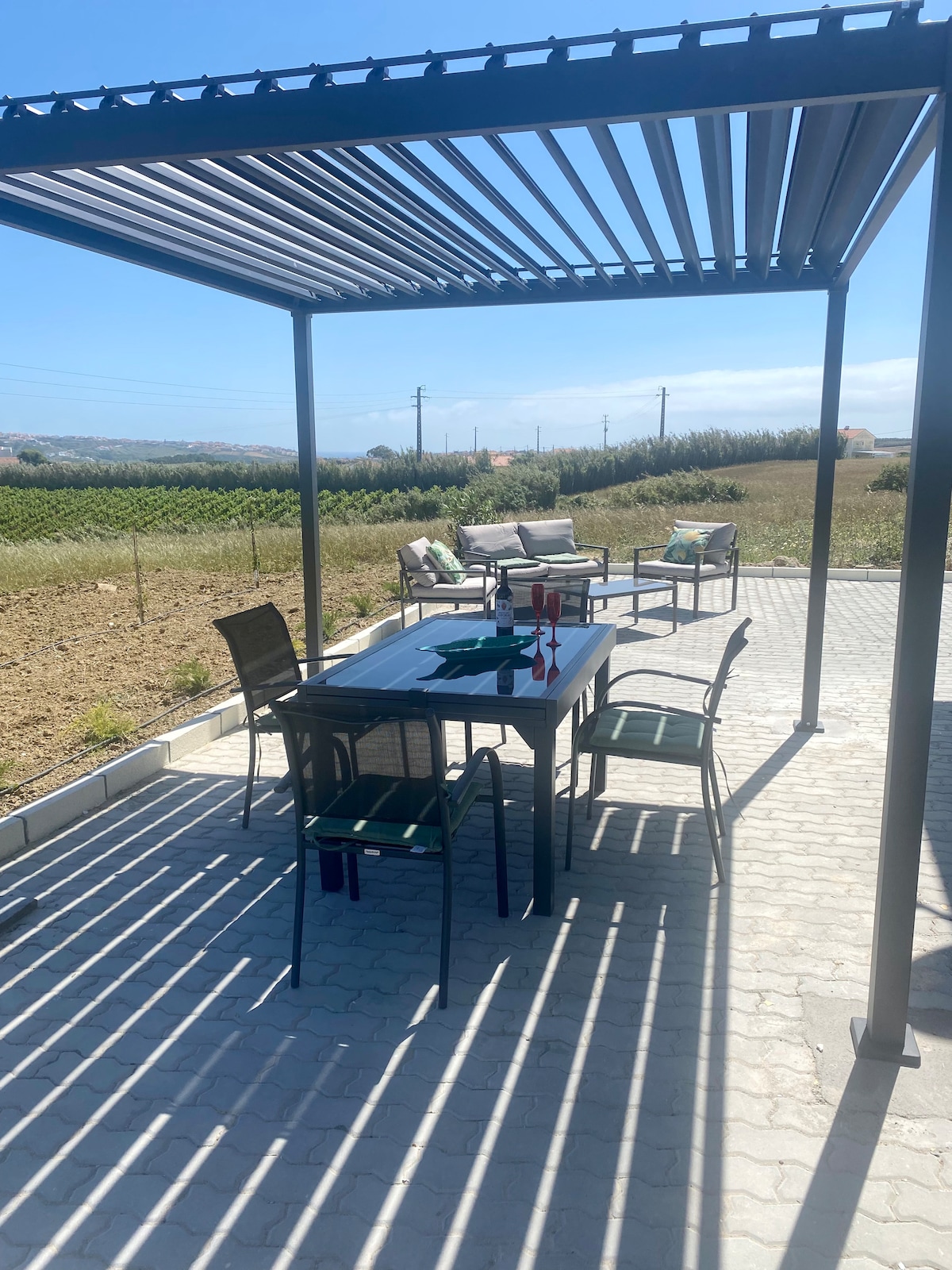 An outdoor dining area is equipped with a large table and several chairs, complemented by a shaded pergola overhead. The surrounding landscape features greenery, providing a backdrop of open space. The setting invites relaxation and dining in nature.