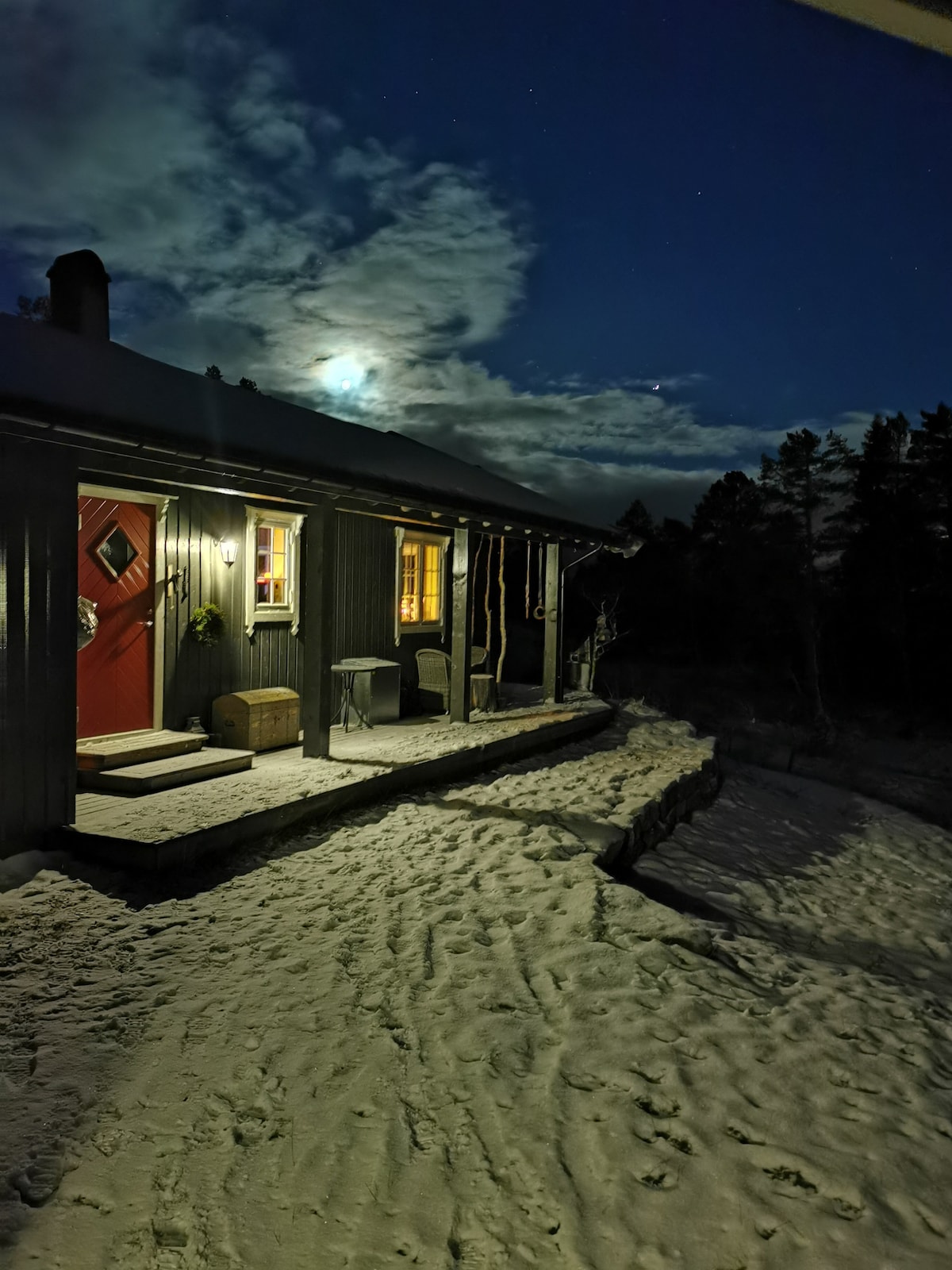 The exterior of the cabin is illuminated by warm light from the windows and the front door, which features a red color. A moonlit sky casts a gentle glow over a snow-covered path leading to the entrance, with trees softly outlined in the background.
