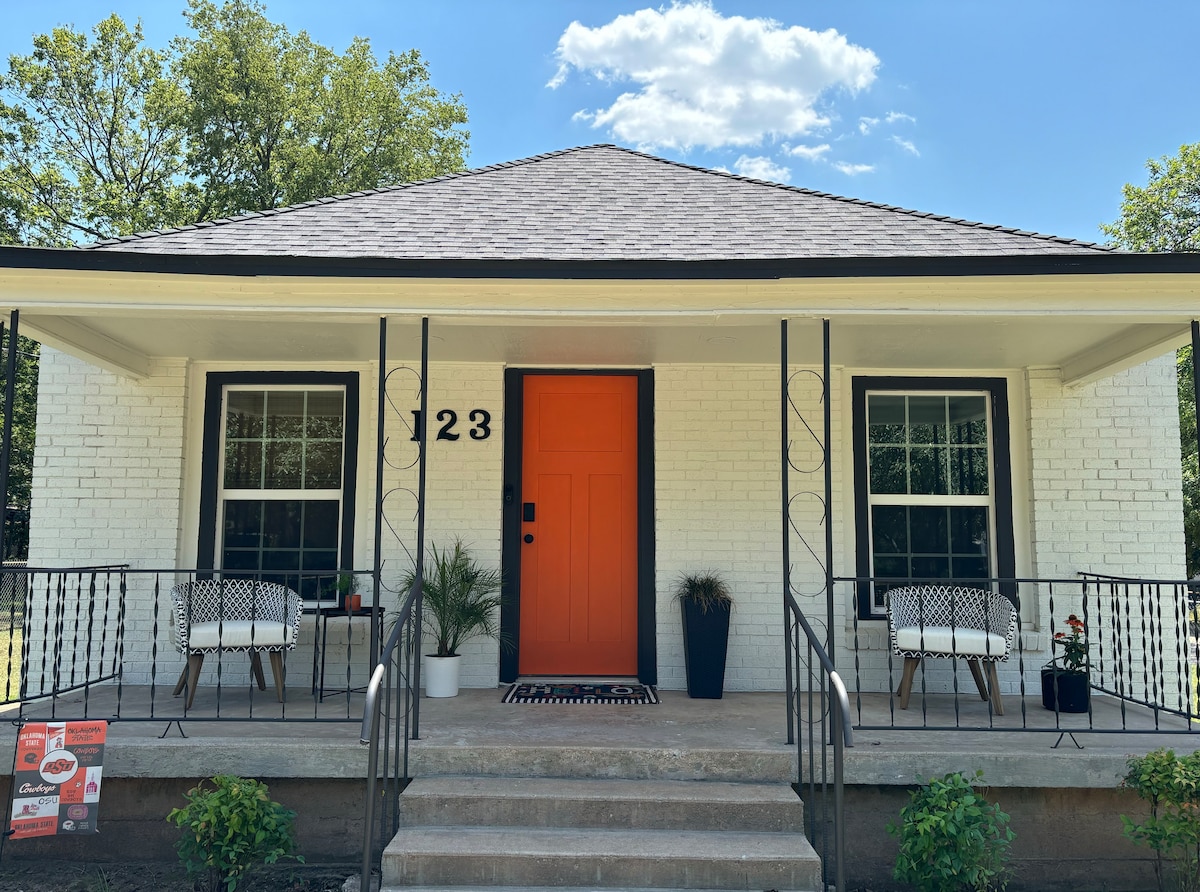 The front porch features a vibrant orange door, flanked by two white chairs and small potted plants. The welcoming entrance is surrounded by large windows and a stylish entryway mat. The house is framed by a lush green landscape under a clear blue sky.