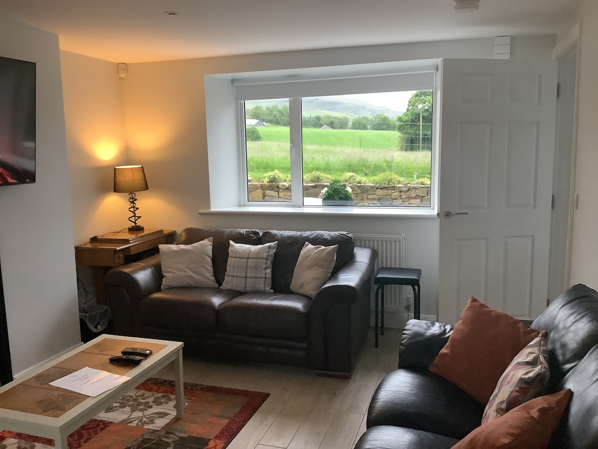 The living area features two comfortable sofas arranged around a low table. Natural light fills the space through a large window, which offers views of the surrounding countryside. A small lamp sits on a side table, adding to the cozy ambiance.