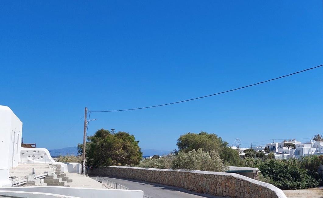 The image captures a clear blue sky over a scenic view featuring a winding road lined with greenery. Whitewashed buildings are visible in the distance, contributing to the serene, coastal landscape.