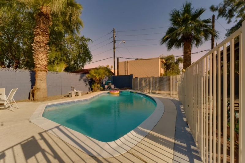 A serene swimming pool is outlined by a white fence, featuring turquoise water reflecting the surrounding palm trees. Lounge chairs are positioned nearby on the sunlit patio, creating a relaxing outdoor space for families. The pool area is tranquil and inviting.