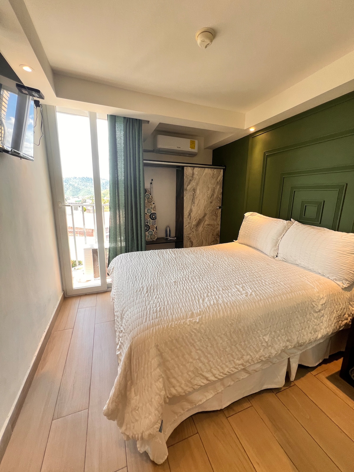 A cozy sleeping area is showcased, featuring a spacious bed with a textured white coverlet. Natural light is allowed in through a large sliding door, complemented by soft green curtains. The room is accented with a green wall and includes a minimalist wardrobe.