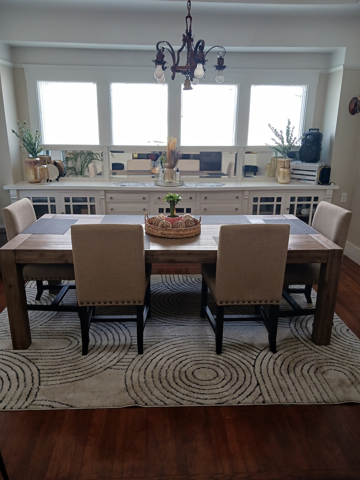 A dining area is presented with a wooden table surrounded by six upholstered chairs. A centerpiece of fresh flowers rests on the table, complemented by a circular rug featuring a spiral design. Bright light enters through large windows, creating an inviting space for gatherings.