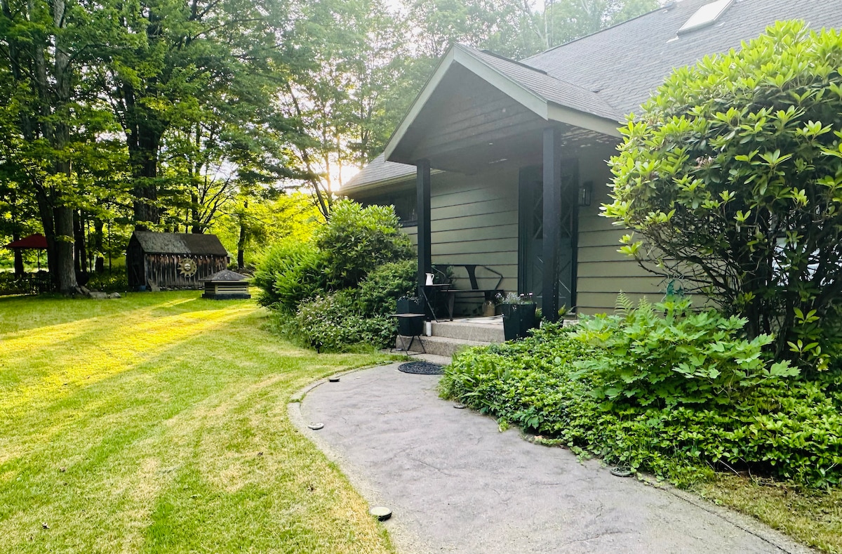 The exterior features a welcoming entrance with stone steps leading to a covered porch. Lush green grass surrounds the home, and a pathway guides to a shed in the background. Natural light filters through the trees, creating a serene outdoor setting.