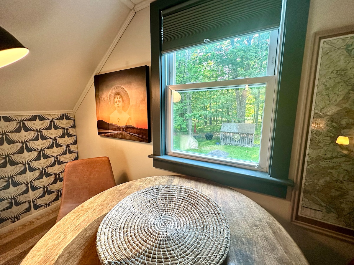 A small dining area is seen with a round wooden table and a patterned chair. A large window offers a view of the outdoors with trees and a rustic shed visible beyond. A piece of artwork is displayed on the wall, enhancing the cozy atmosphere.