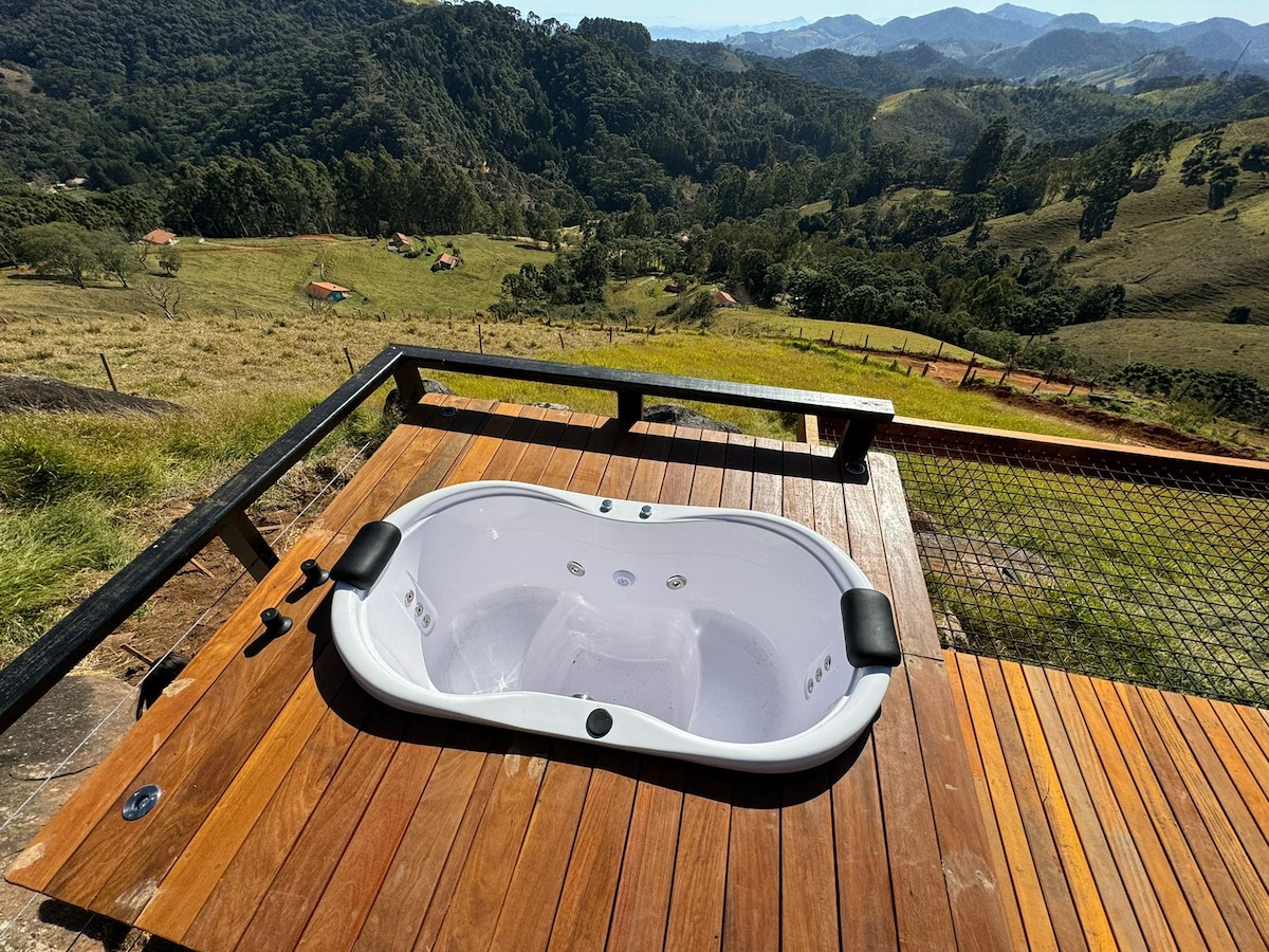 A double hot tub is situated on a wooden deck, overlooking a stunning vista of rolling hills and valleys. The landscape is lush, featuring various shades of green, with distant mountains creating a serene backdrop.