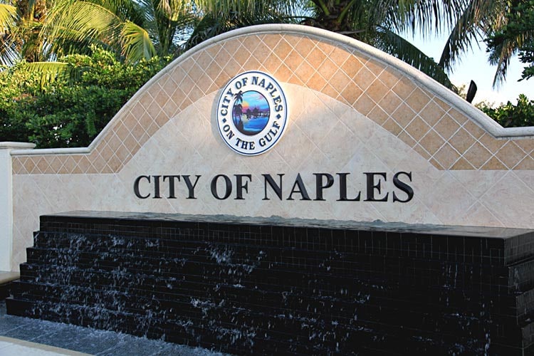 A decorative sign for the City of Naples is displayed against a tiled backdrop. Water cascades over a black stone feature below the sign, framed by lush palm trees. The sign includes the city logo and a subtitle highlighting Naples as 'On the Gulf.'