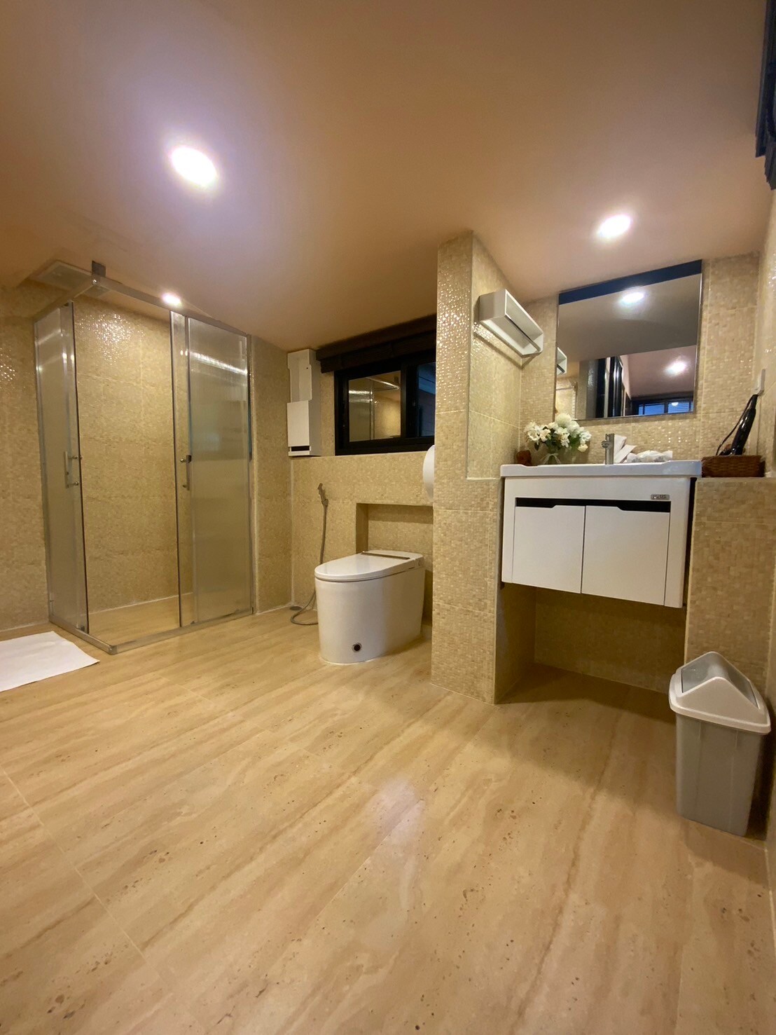 A spacious bathroom is fitted with modern amenities, including a walk-in shower with glass doors and elegant tile work. A white toilet and a sleek vanity with storage complement the design. Natural light enters through a window, enhancing the bright and clean setting.