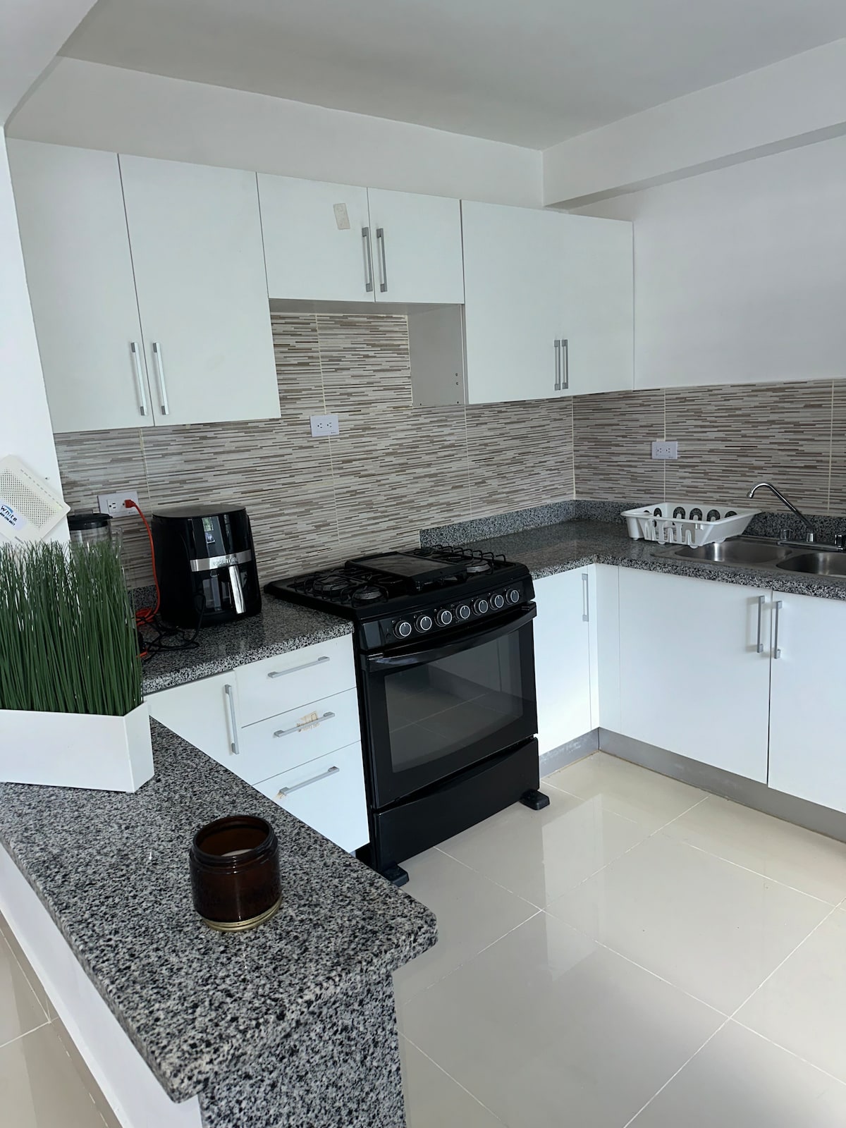 A modern kitchen is presented with sleek white cabinetry and a gray granite countertop. A black stove is positioned against the wall, complemented by a stainless steel sink and an array of appliances. Light reflects softly off the tile flooring, enhancing the clean, contemporary aesthetic.