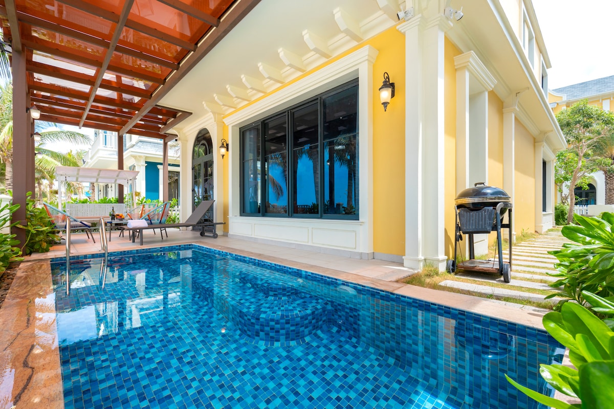 A private terrace features a glistening swimming pool surrounded by lush greenery. Floor-to-ceiling windows provide a view into the living area. Comfortable loungers and a barbecue grill are positioned nearby, creating an inviting outdoor space for relaxation and leisure.