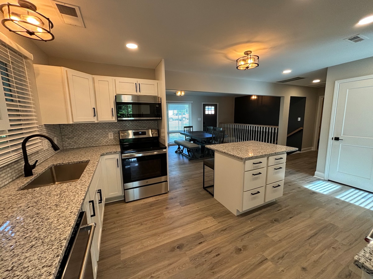 The kitchen features modern stainless steel appliances and a spacious countertop with a light granite finish. An island in the center provides additional workspace. Natural light flows through the nearby windows, enhancing the open layout that connects to the dining area.
