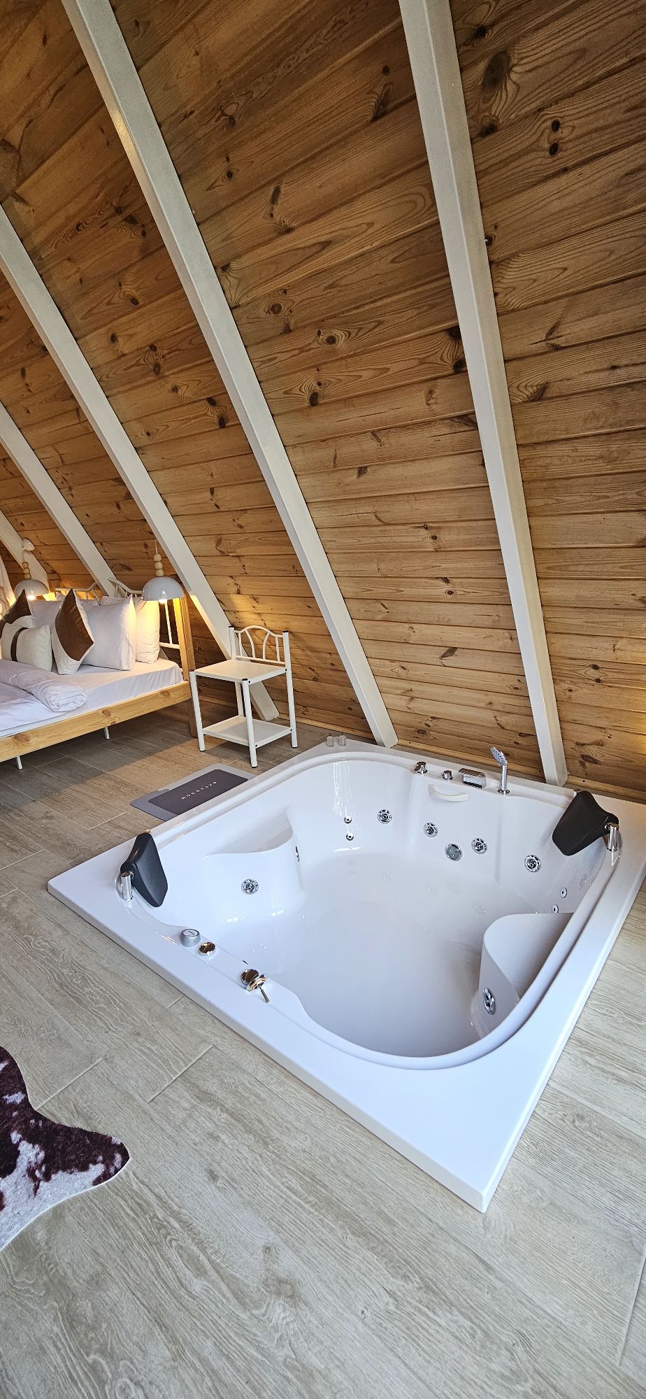 A private jetted tub is positioned on a light tiled floor, accompanied by wooden walls that add warmth to the space. A cozy sleeping area with a bed is visible in the background, creating a serene atmosphere for relaxation.