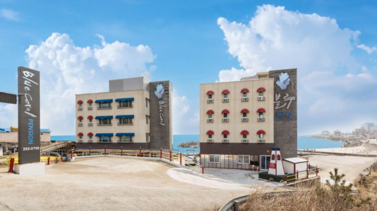 The exterior of the Blue Coral Pension is presented, featuring a modern building with multiple floors and distinctive red awnings above the windows. A wide parking area is visible in front, with a clear view of the ocean in the background under a bright blue sky.