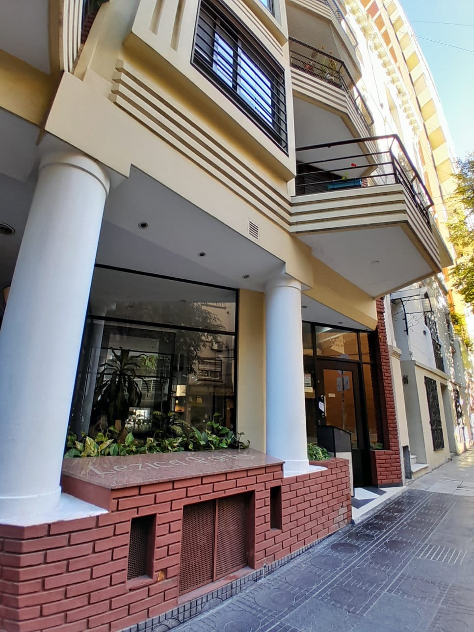 The exterior of the building features prominent white columns and large glass windows, allowing natural light to illuminate the interior. Green plants are arranged in a planter near the entrance, while a wide sidewalk provides easy access to the main entrance.