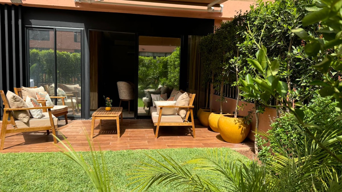 A garden area features wooden decking, stylish outdoor seating, and a coffee table. Lush greenery surrounds the space, while vibrant planters add a pop of color. Large glass doors lead into the interior, seamlessly connecting the outdoor and indoor environments.