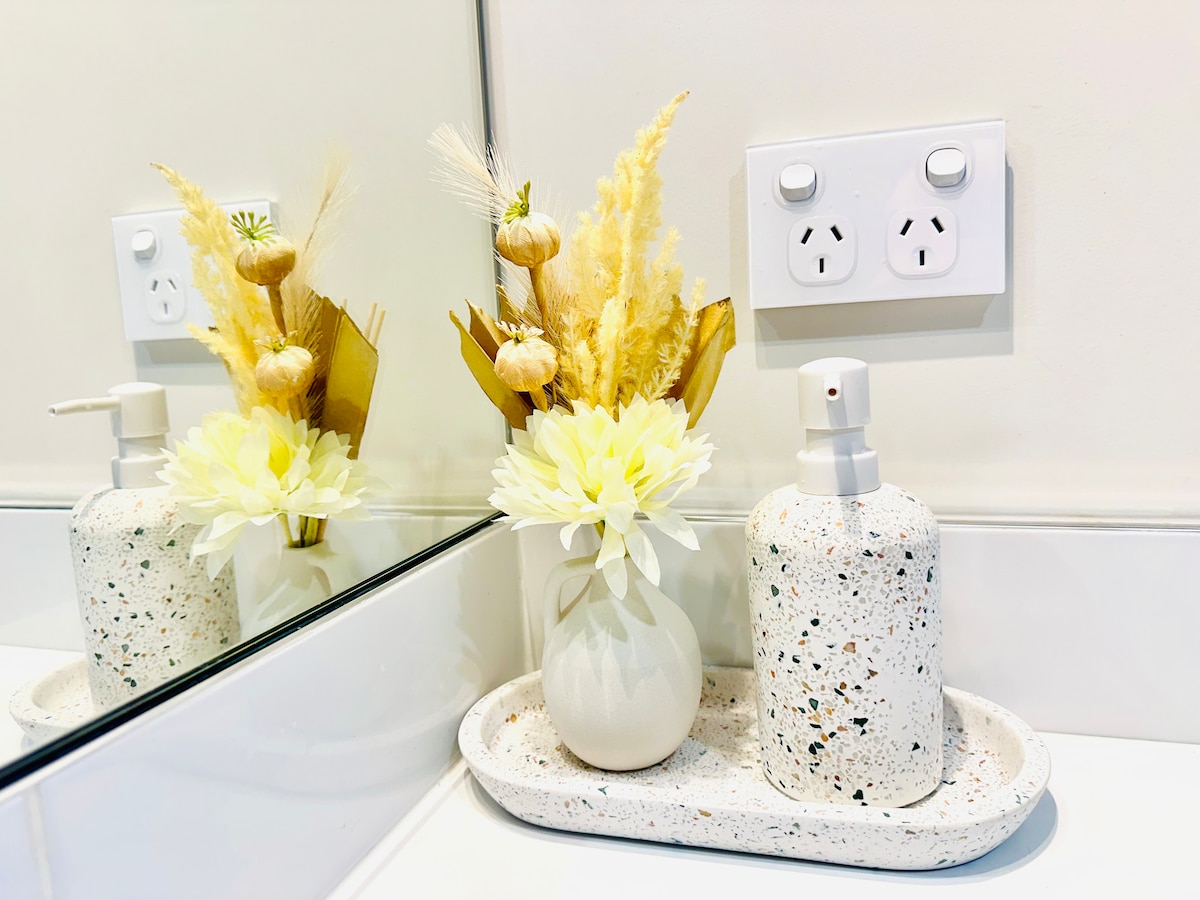 A bathroom countertop is shown featuring a decorative arrangement of dried flowers in a light-colored vase. A speckled soap dispenser sits alongside a textured serving tray, all against a backdrop of a mirror and electrical outlets.