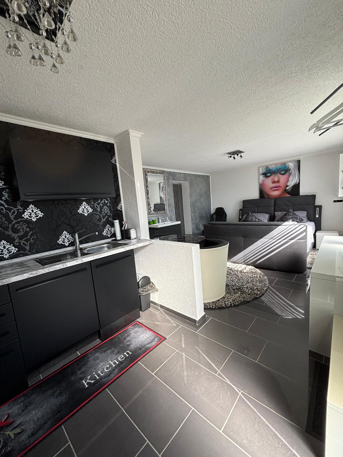 A modern kitchen area features sleek black cabinetry and a marble countertop, equipped with a television mounted above. An inviting living space is visible in the background, showcasing a bed with dark bedding and contemporary decor. Large windows allow for natural light to illuminate the room.