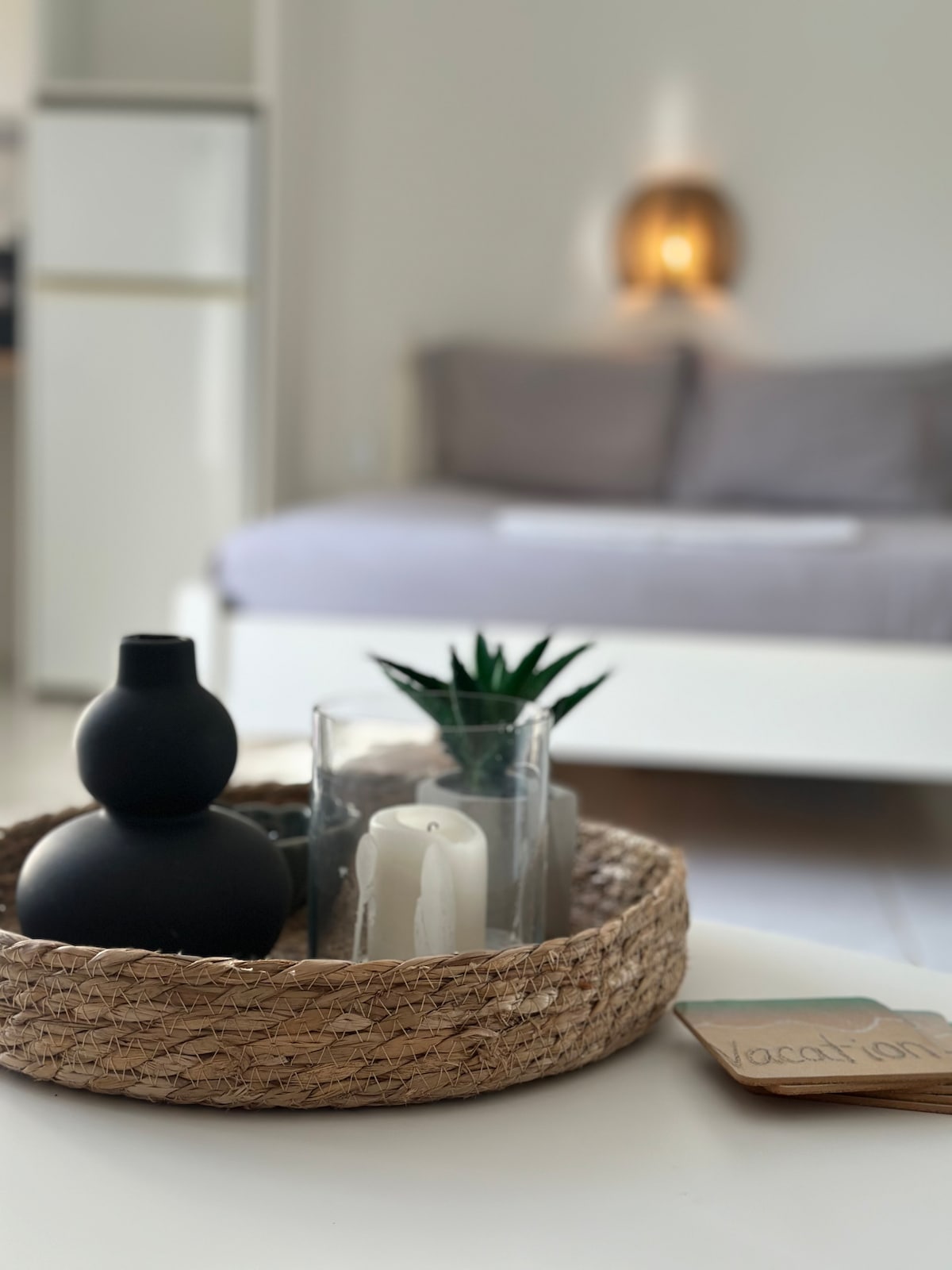 A decorative table setting features a woven basket containing a black vase, white candles, and a small green plant. In the background, a softly lit area showcases a spacious bed with neutral-colored bedding, promoting a calm ambiance.