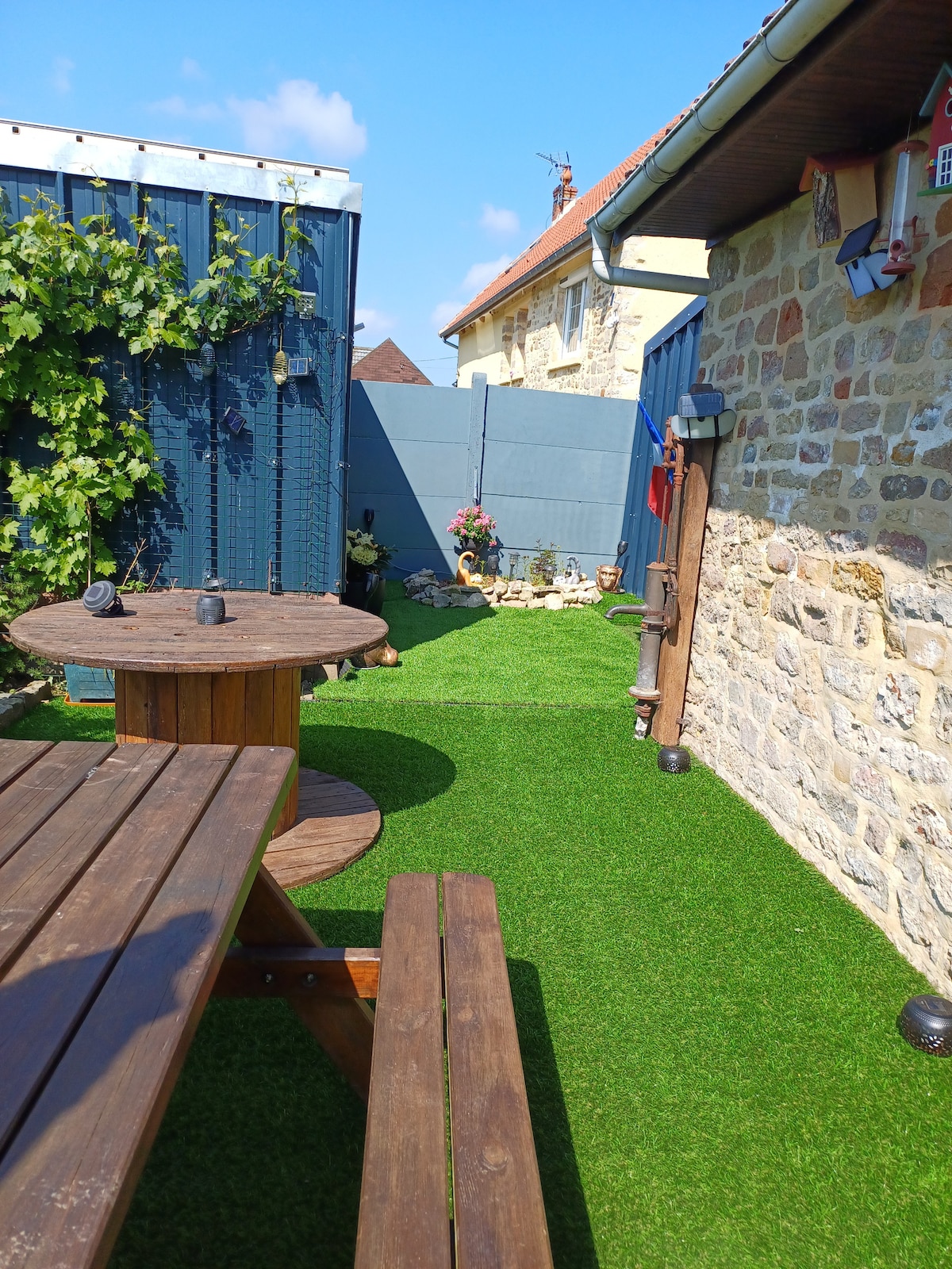 An outdoor space features artificial grass and a wooden picnic table with attached benches. A stone wall and blue fencing provide a backdrop, while decorative elements like flowers and garden ornaments are seen throughout the area, creating a pleasant and serene environment.