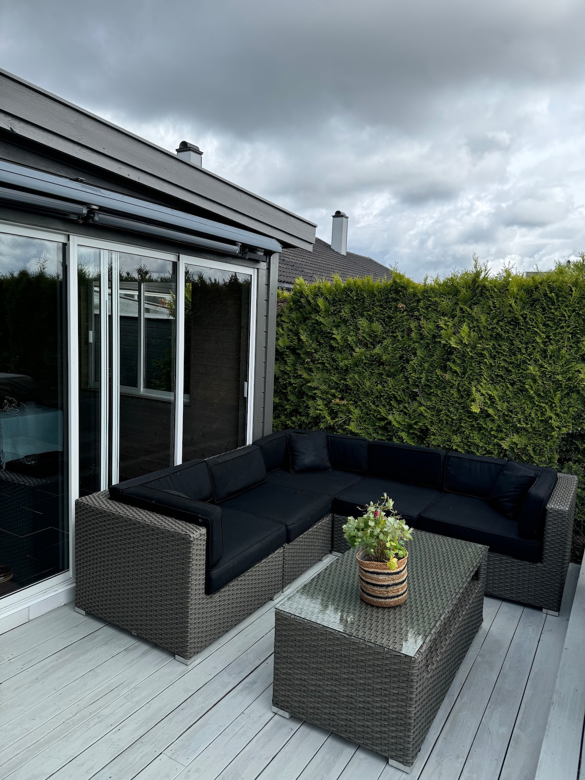 An outdoor seating area features a dark wicker sectional couch and a glass-topped coffee table. A potted plant decorates the table. Green hedges provide privacy, and large glass doors offer easy access to the indoor space.